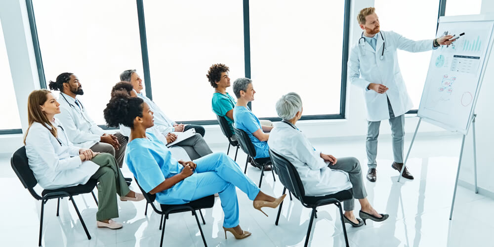 A nurse educator stands at the front of a classroom, in front of a group of nurses.